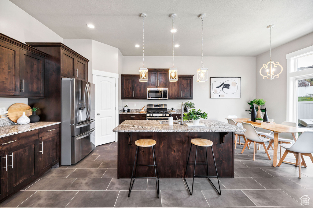 Kitchen with dark brown cabinetry, light stone counters, a kitchen island with sink, stainless steel appliances, and a kitchen bar