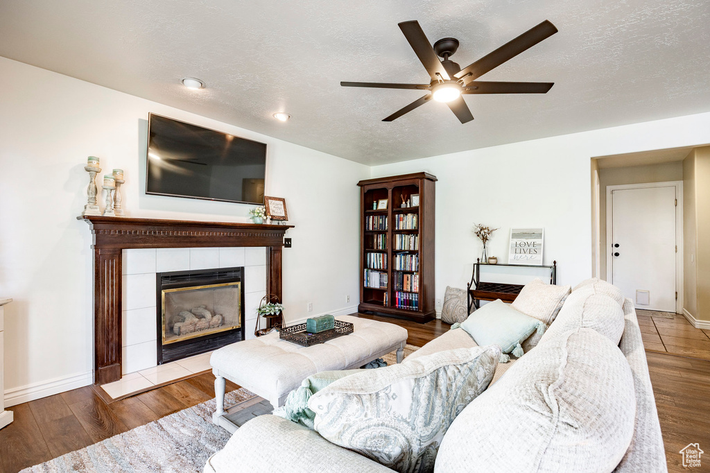 Living room with a tiled fireplace, a ceiling fan, wood finished floors, baseboards, and a textured ceiling