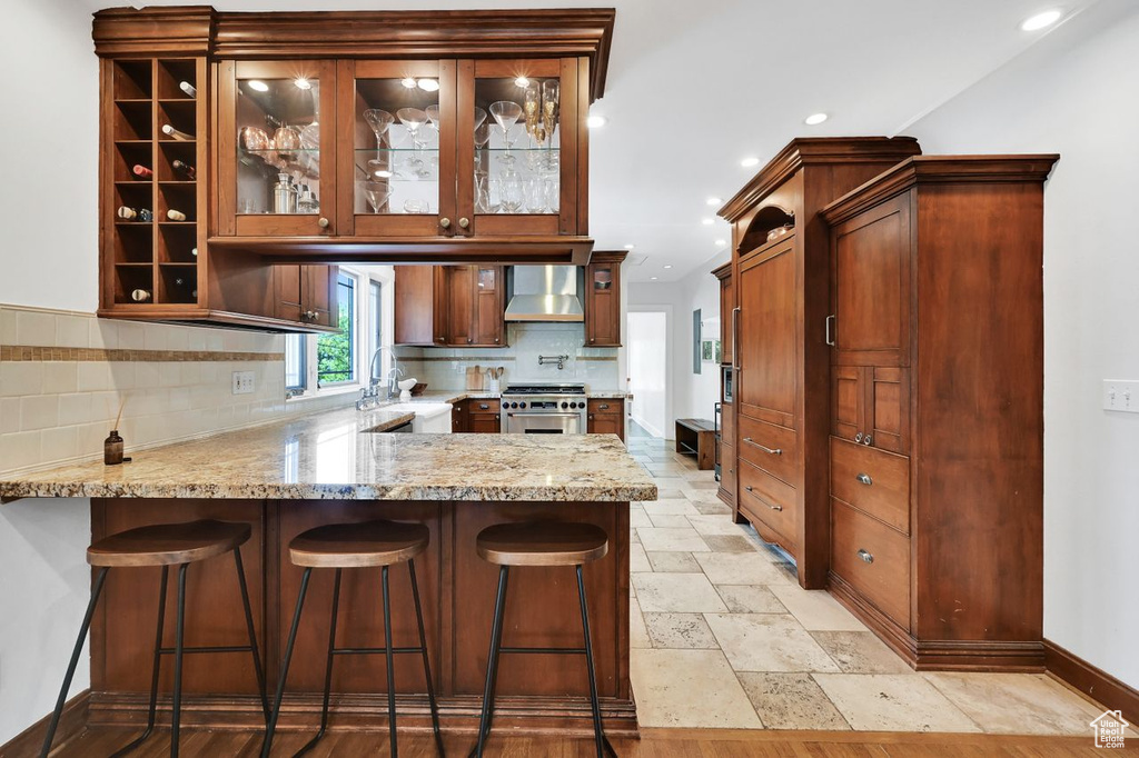 Kitchen with wall chimney range hood, stainless steel stove, a sink, a peninsula, and glass insert cabinets