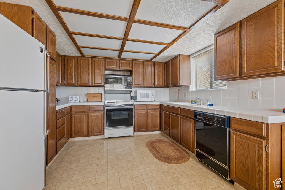 Kitchen with brown cabinetry, white appliances, tile counters, a sink, and backsplash