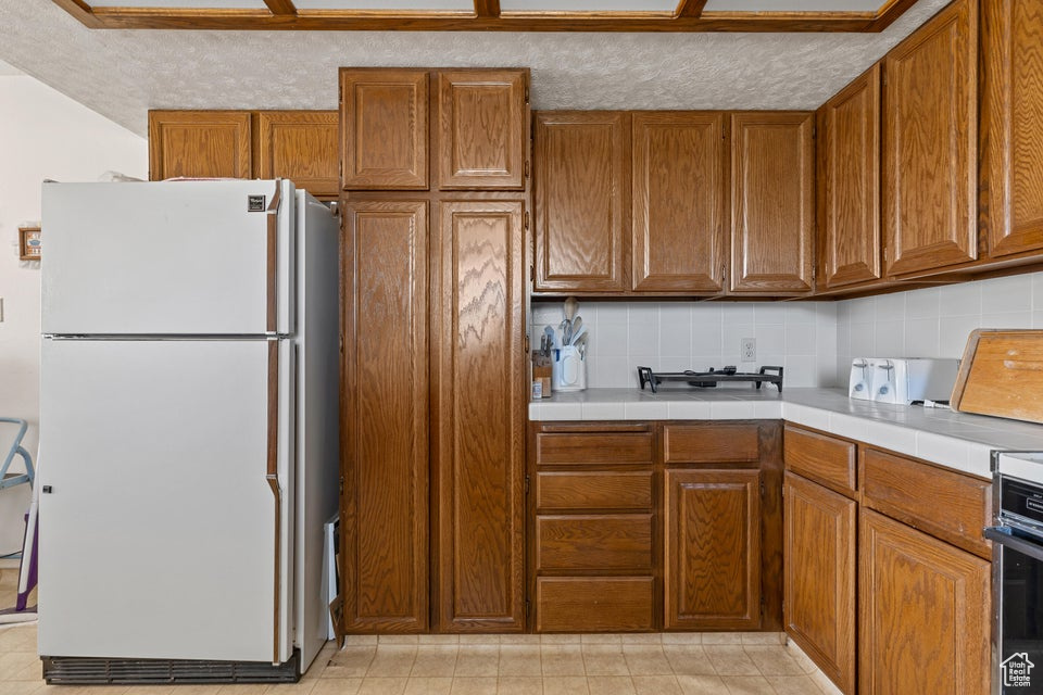 Kitchen featuring decorative backsplash, freestanding refrigerator, brown cabinetry, and tile counters