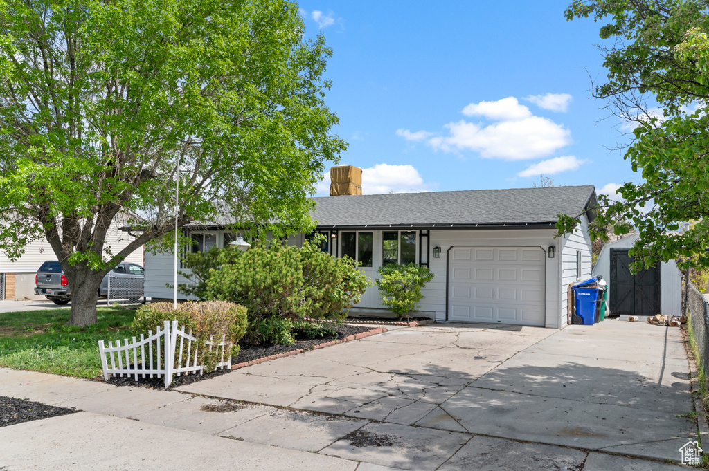 Single story home featuring driveway, a chimney, a shingled roof, an attached garage, and a storage shed