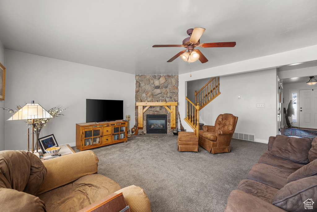 Carpeted living area featuring ceiling fan, a stone fireplace, visible vents, stairway, and baseboards