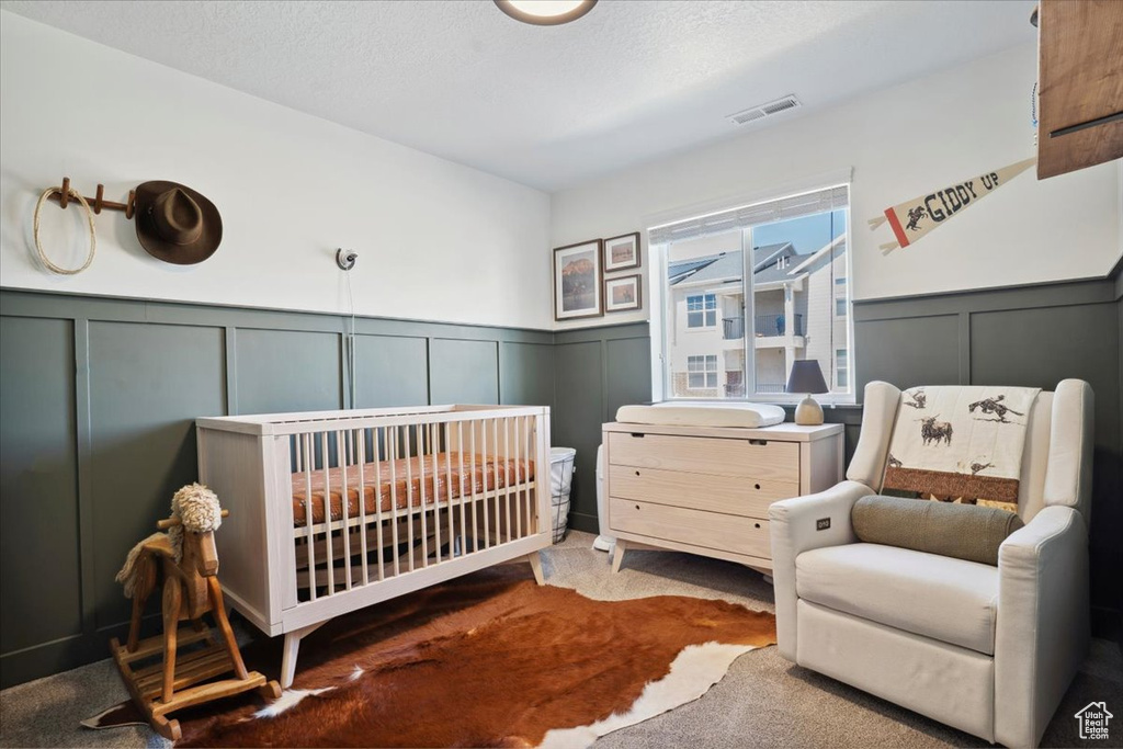 Carpeted bedroom with visible vents, a decorative wall, a nursery area, and a wainscoted wall