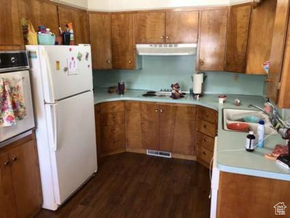 Kitchen with brown cabinets, white appliances, under cabinet range hood, visible vents, and dark wood-style floors