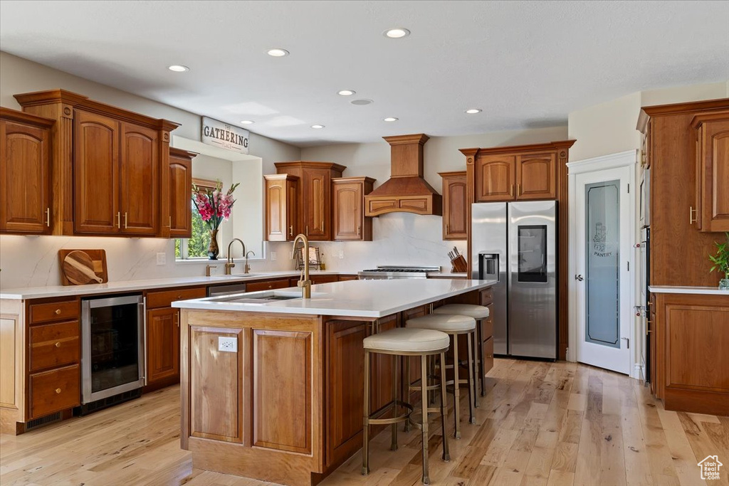 Kitchen featuring stainless steel fridge, beverage cooler, an island with sink, premium range hood, and recessed lighting