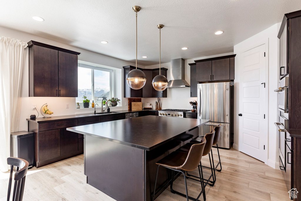 Kitchen with dark brown cabinetry, light wood-style floors, a breakfast bar area, wall chimney exhaust hood, and stainless steel appliances