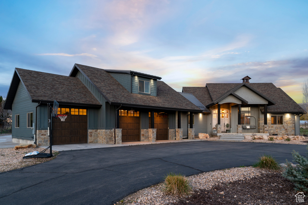 View of front facade with driveway, stone siding, a shingled roof, covered porch, and an attached garage