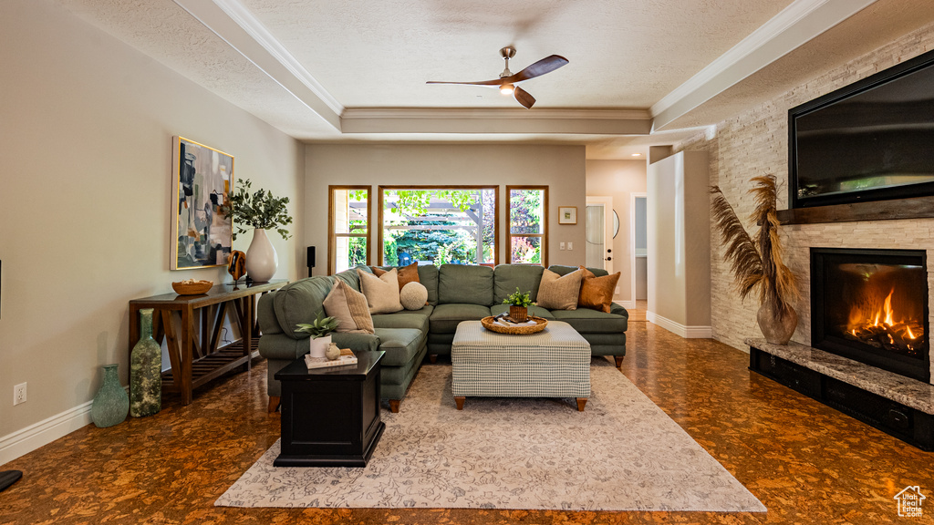Living area featuring a tray ceiling, a ceiling fan, ornamental molding, a stone fireplace, and a textured ceiling