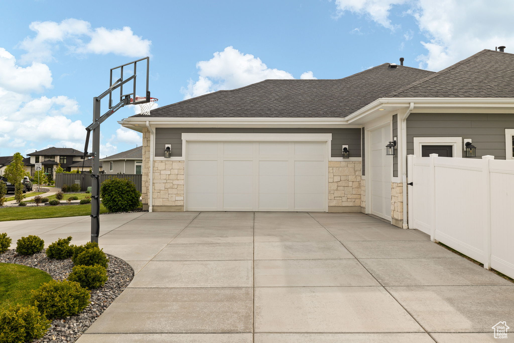View of property exterior featuring fence, a shingled roof, concrete driveway, and stone siding