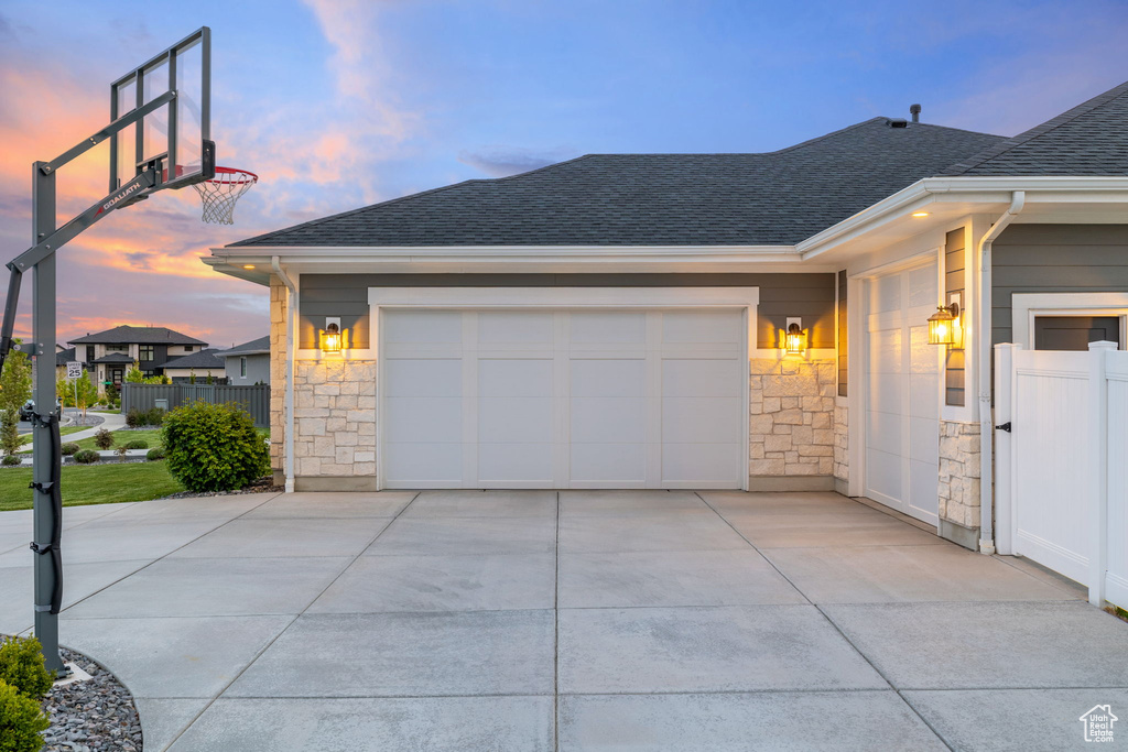Property exterior at dusk featuring fence, stone siding, driveway, roof with shingles, and an attached garage