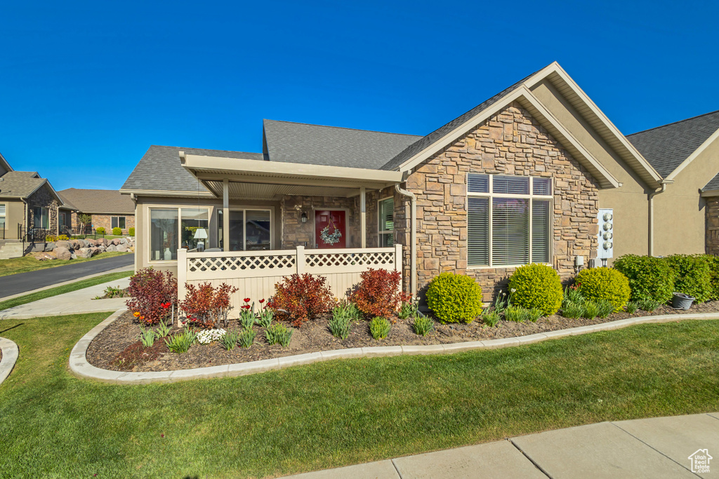 Ranch-style home featuring covered porch, roof with shingles, a front yard, and stone siding