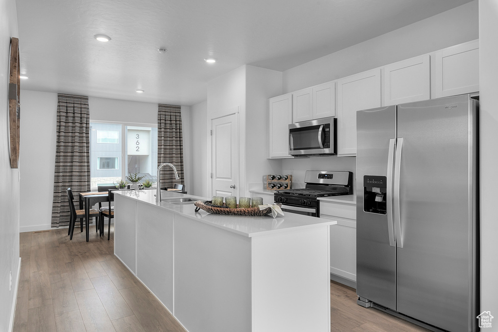 Kitchen with appliances with stainless steel finishes, a sink, light wood finished floors, a kitchen island with sink, and white cabinetry