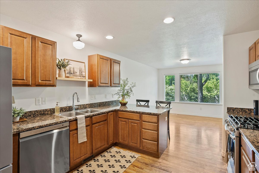 Kitchen with appliances with stainless steel finishes, a sink, a peninsula, light wood-style flooring, and brown cabinetry