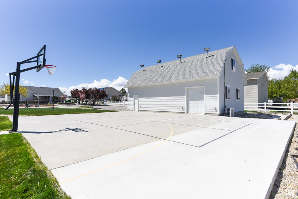View of side of property featuring fence, community basketball court, a detached garage, and roof with shingles