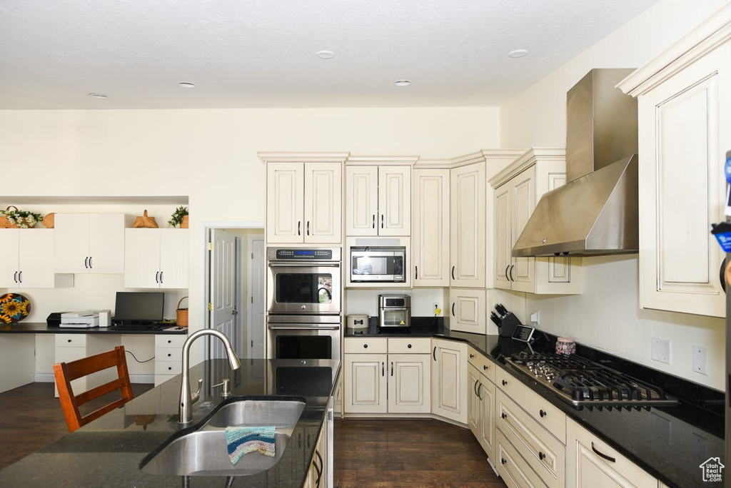 Kitchen with appliances with stainless steel finishes, a sink, wall chimney exhaust hood, dark stone countertops, and dark wood-style floors