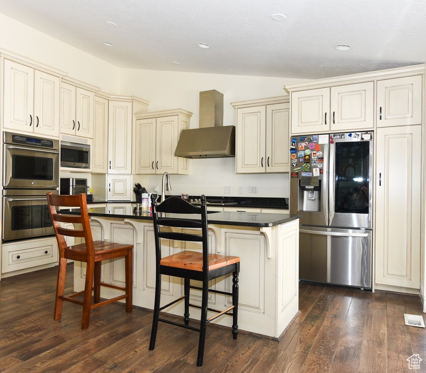 Kitchen with a kitchen bar, dark countertops, stainless steel appliances, wall chimney exhaust hood, and cream cabinetry