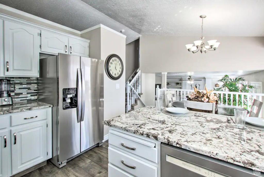 Kitchen with light stone counters, ceiling fan with notable chandelier, decorative backsplash, appliances with stainless steel finishes, and dark wood-style floors