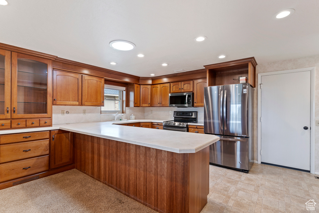 Kitchen featuring a peninsula, glass insert cabinets, light countertops, brown cabinets, and stainless steel appliances
