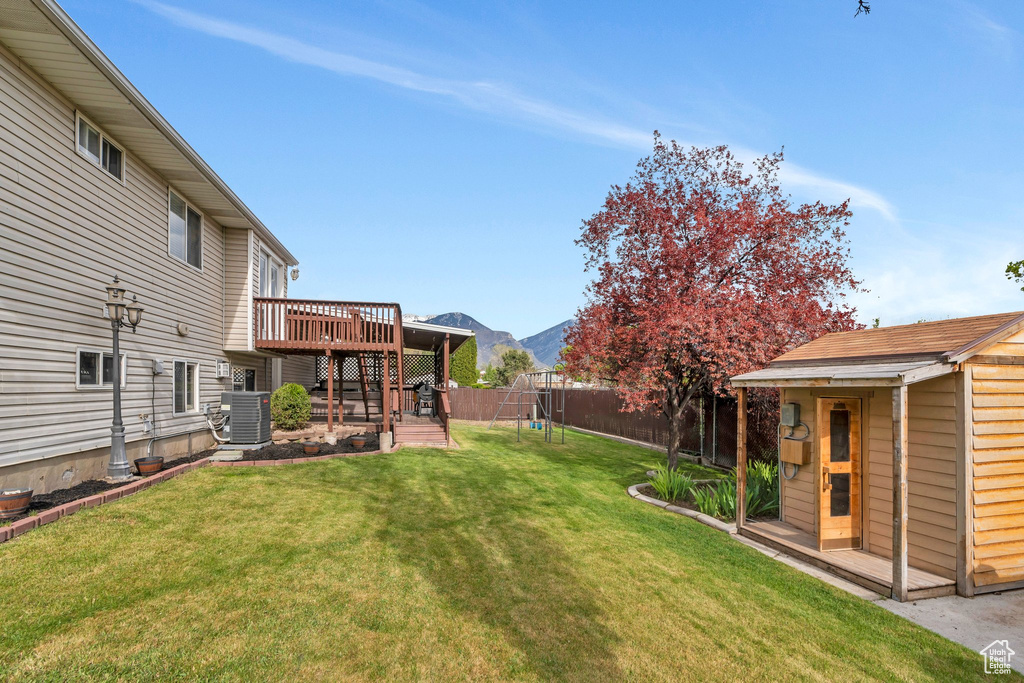 View of yard with a fenced backyard, a deck with mountain view, central AC unit, a playground, and an outbuilding