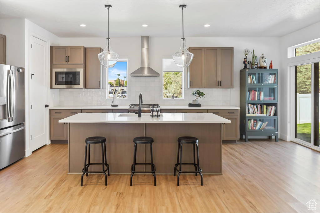 Kitchen with light wood-type flooring, wall chimney exhaust hood, appliances with stainless steel finishes, and tasteful backsplash