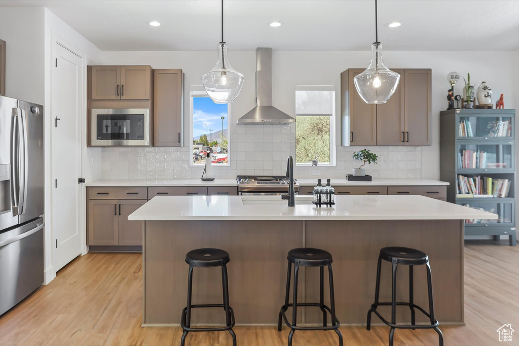 Kitchen with wall chimney range hood, appliances with stainless steel finishes, light wood finished floors, light countertops, and decorative backsplash