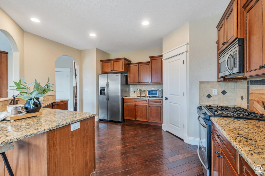 Kitchen featuring dark wood-style floors, brown cabinets, light stone counters, arched walkways, and appliances with stainless steel finishes