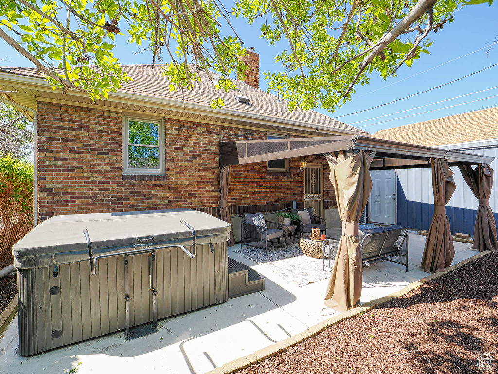 Rear view of property with a chimney, roof with shingles, a hot tub, brick siding, and a patio