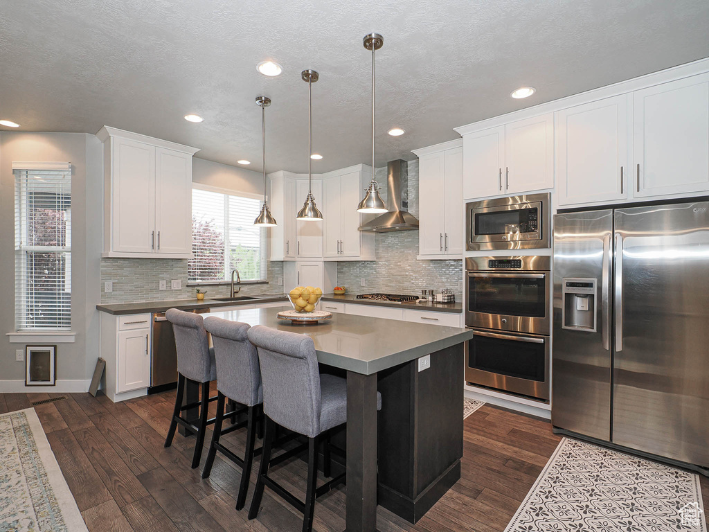 Kitchen with wall chimney range hood, white cabinets, appliances with stainless steel finishes, a sink, and dark wood-style floors