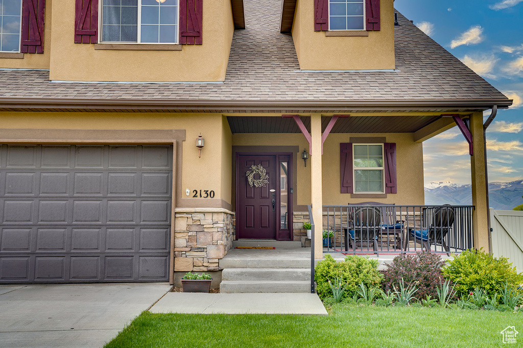 View of exterior entry with roof with shingles, stucco siding, a porch, and stone siding