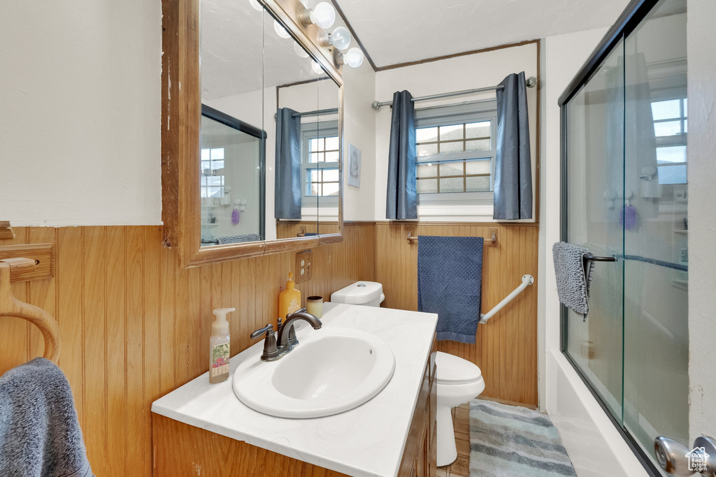 Bathroom with wainscoting, vanity, toilet, wood walls, and bath / shower combo with glass door