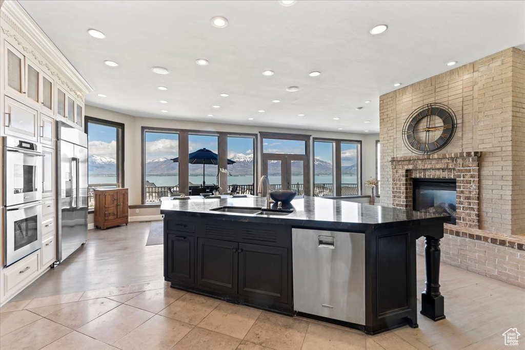 Kitchen featuring a kitchen island with sink, a fireplace, dark cabinetry, a sink, and stainless steel appliances