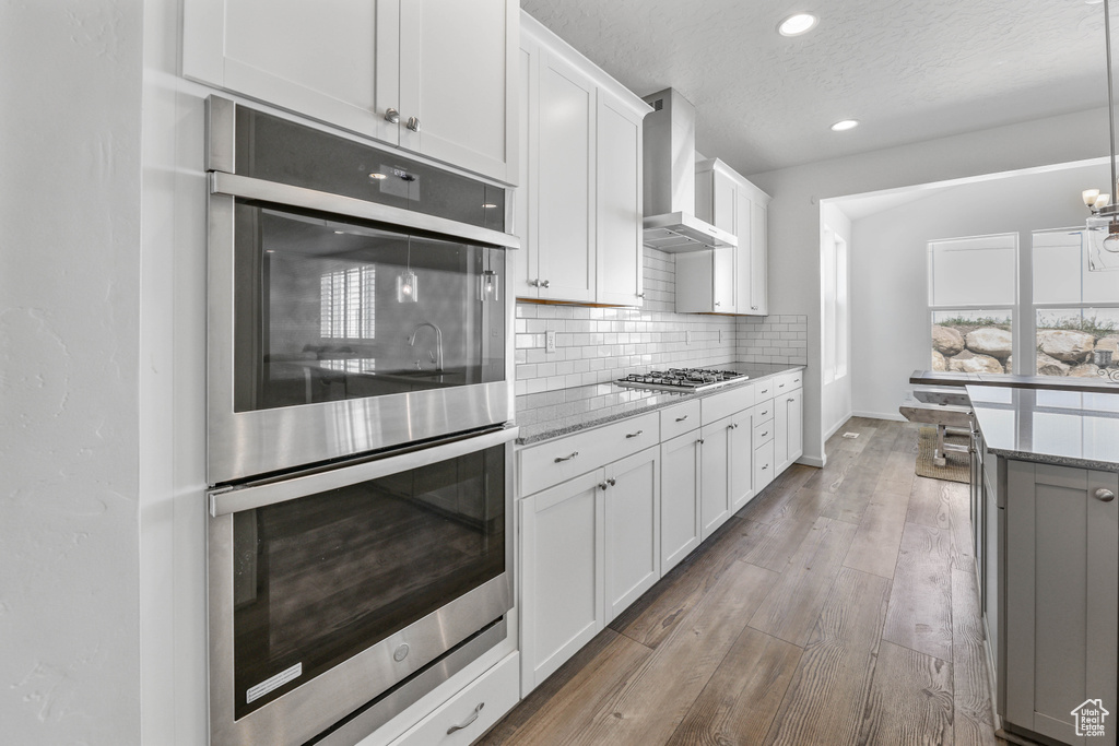 Kitchen featuring light stone countertops, wall chimney exhaust hood, stainless steel appliances, white cabinetry, and decorative backsplash