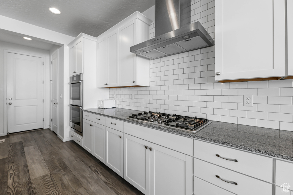 Kitchen featuring wall chimney range hood, white cabinets, dark wood finished floors, dark stone counters, and stainless steel appliances