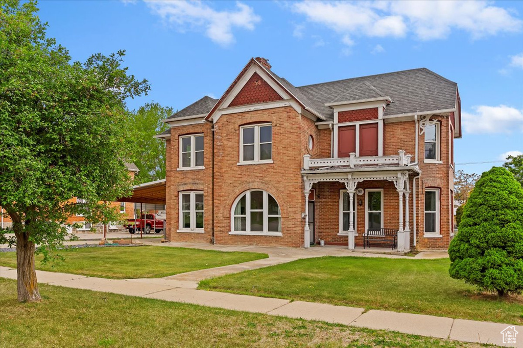 View of front of home with a balcony, a front yard, roof with shingles, brick siding, and a porch