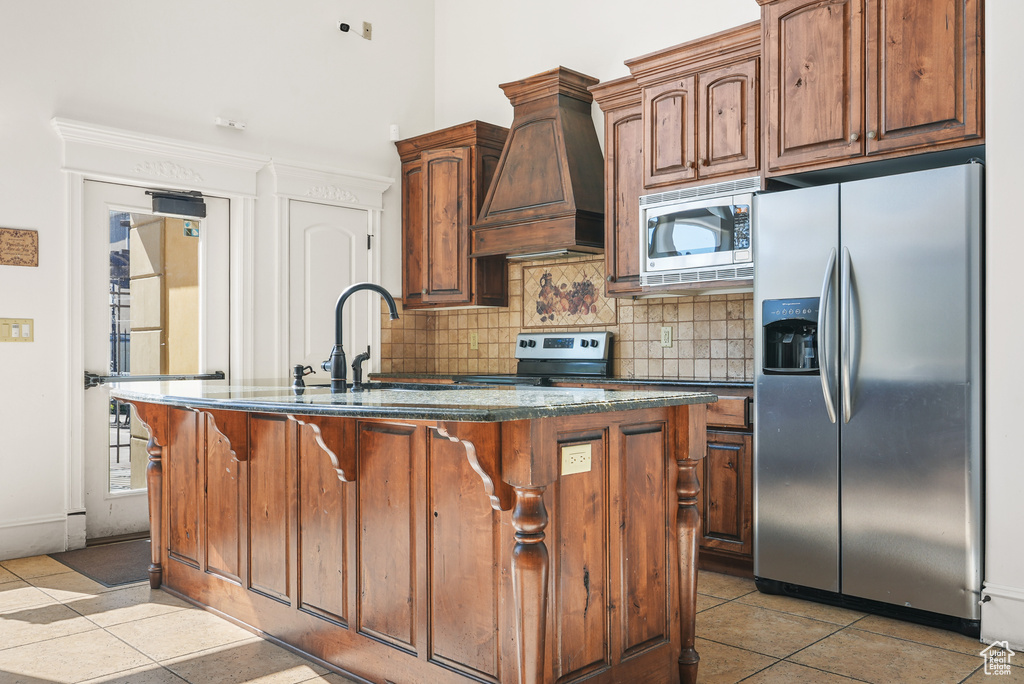 Kitchen featuring custom exhaust hood, light tile patterned flooring, a center island with sink, appliances with stainless steel finishes, and backsplash