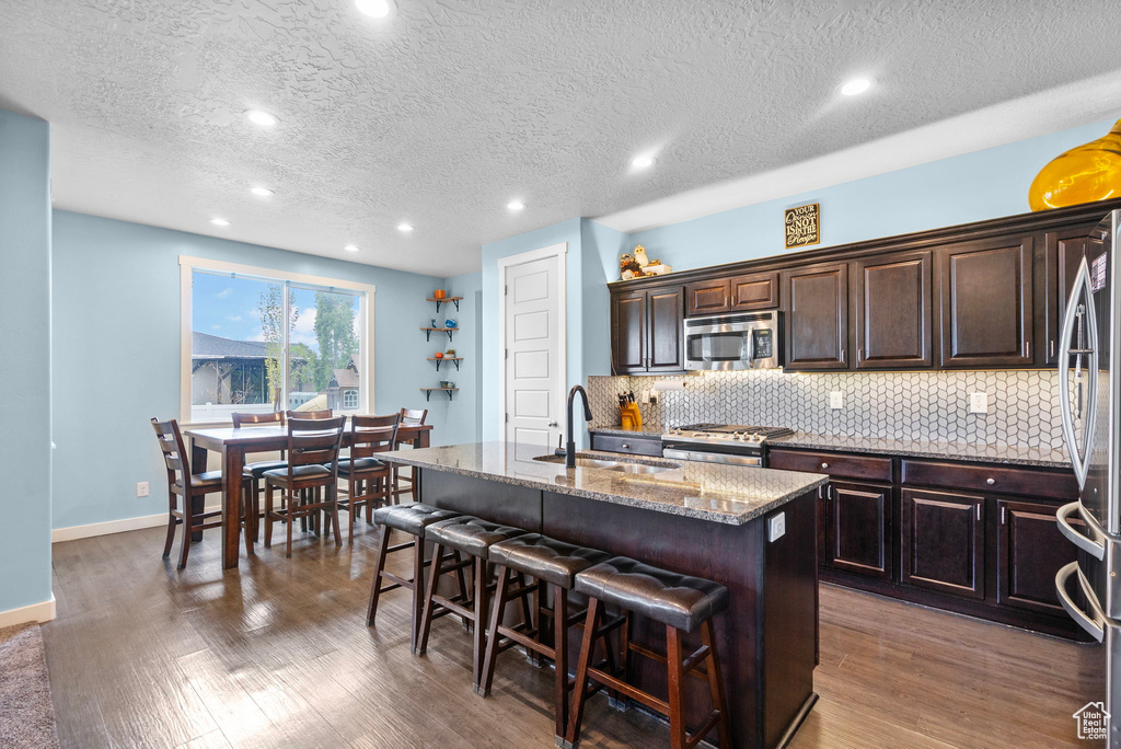 Kitchen with a sink, an island with sink, stainless steel appliances, wood finished floors, and tasteful backsplash