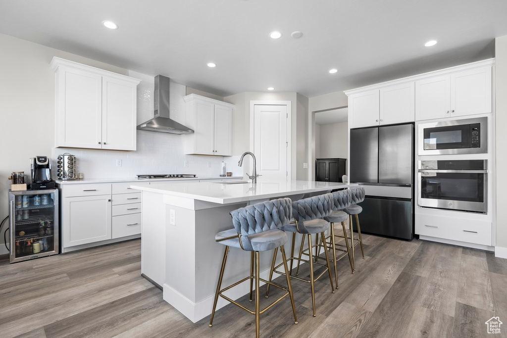 Kitchen featuring a kitchen island with sink, light wood-style floors, wall chimney range hood, a sink, and appliances with stainless steel finishes