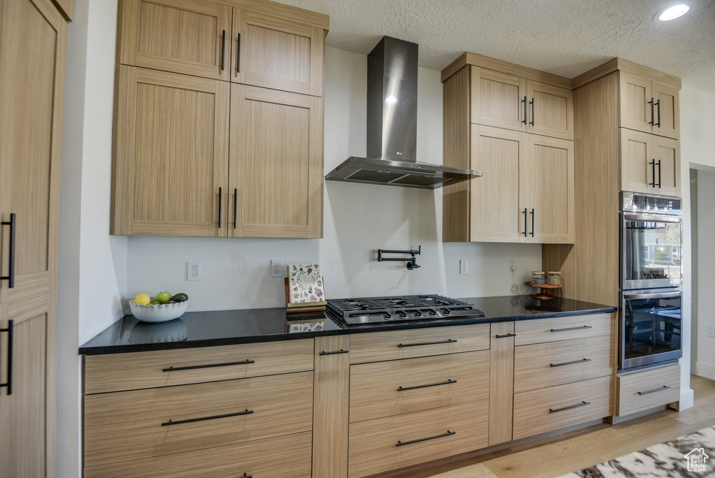 Kitchen featuring dark countertops, light brown cabinetry, stainless steel appliances, and wall chimney exhaust hood