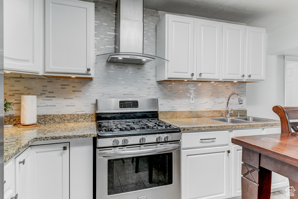 Kitchen with wall chimney range hood, gas stove, a sink, and white cabinetry