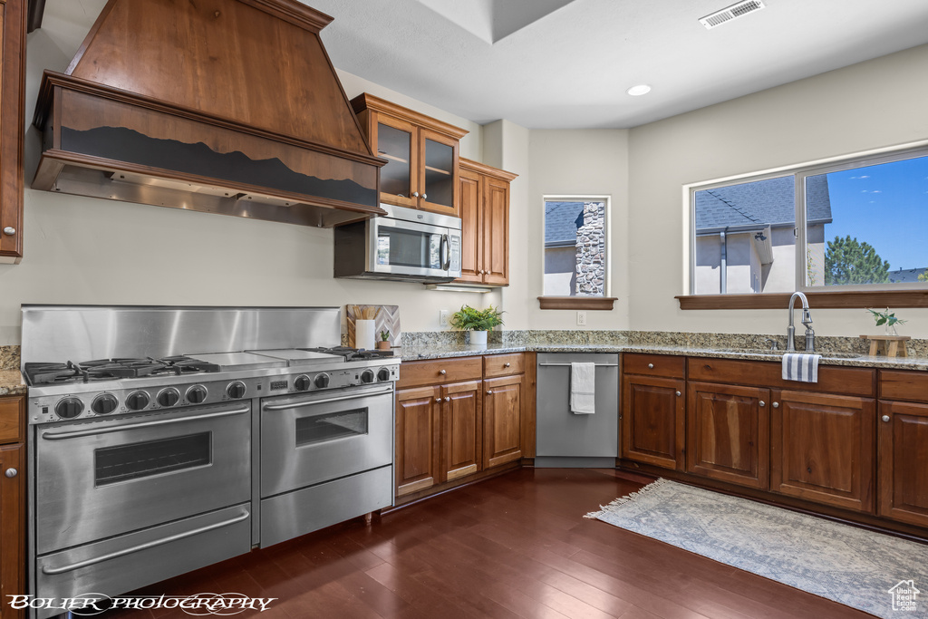 Kitchen featuring appliances with stainless steel finishes, light stone countertops, a sink, dark wood-style floors, and custom range hood
