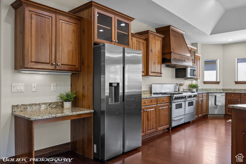 Kitchen with dark wood-style flooring, appliances with stainless steel finishes, brown cabinets, glass insert cabinets, and custom range hood