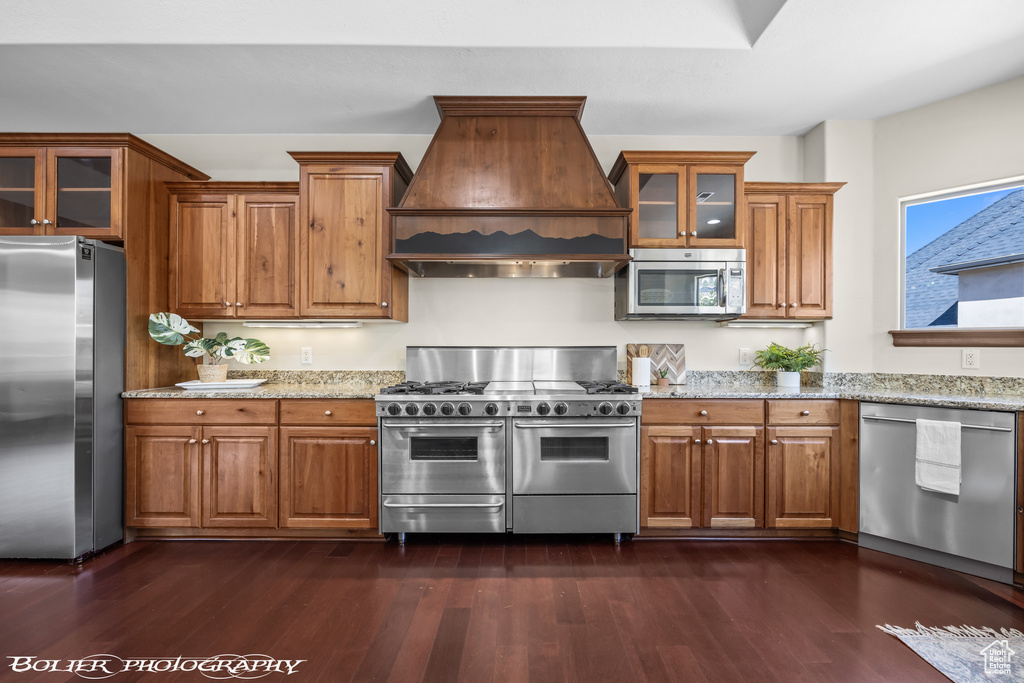 Kitchen featuring brown cabinetry, light stone countertops, glass insert cabinets, premium range hood, and stainless steel appliances