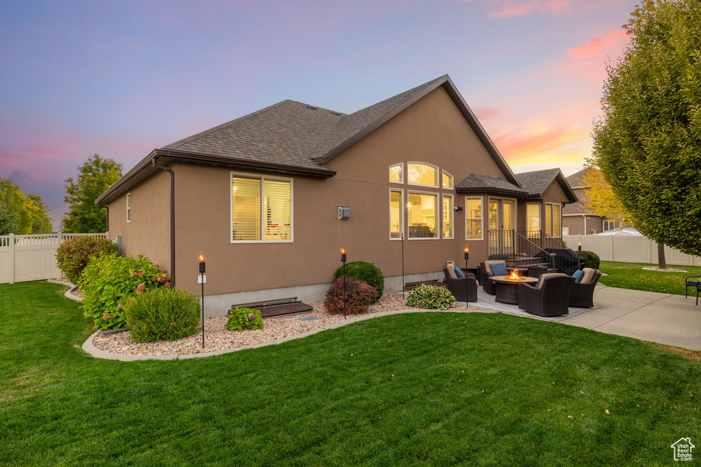 Back of house at dusk with a fenced backyard, a shingled roof, a patio area, a lawn, and a fire pit