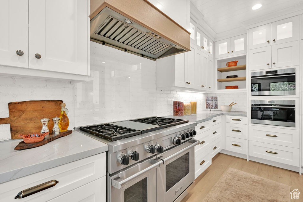 Kitchen with decorative backsplash, custom exhaust hood, light stone countertops, white cabinetry, and range with two ovens
