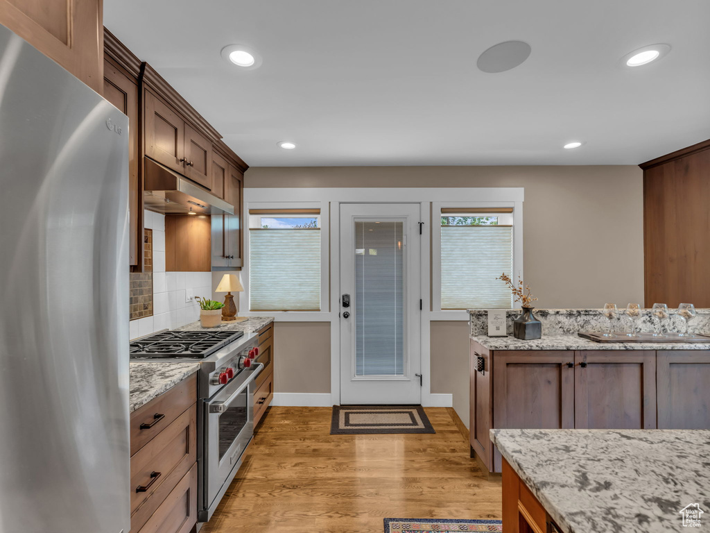 Kitchen with decorative backsplash, light wood-style floors, under cabinet range hood, light stone counters, and appliances with stainless steel finishes