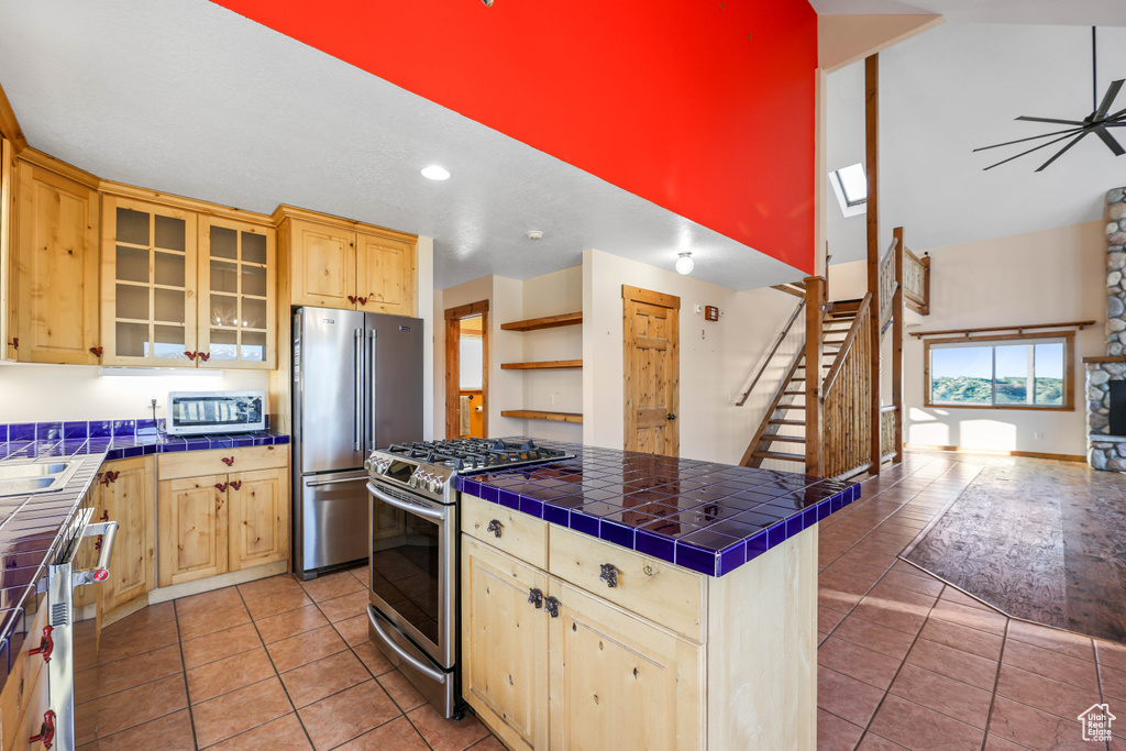 Kitchen featuring tile counters, a towering ceiling, light brown cabinetry, and stainless steel appliances