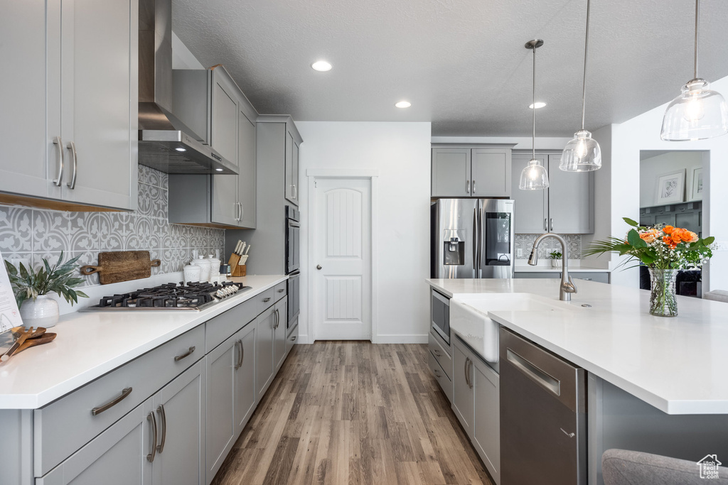 Kitchen with wood finished floors, gray cabinetry, a sink, stainless steel appliances, and wall chimney exhaust hood