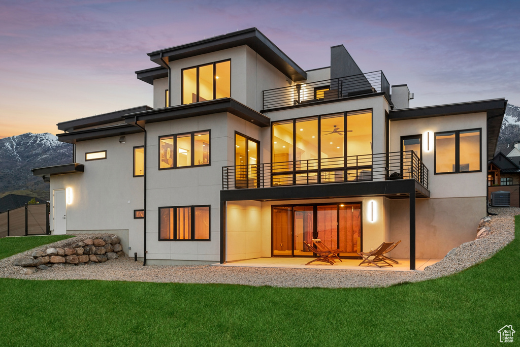 Back of property at dusk featuring a lawn, central AC unit, a patio area, a balcony, and stucco siding