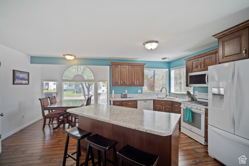 Kitchen featuring white appliances, a sink, dark wood-style floors, a center island, and a kitchen breakfast bar